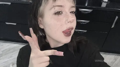 VernellZiccardi sticks out her tongue while wearing a black shirt and showing off her long, pink-tipped nails and nose ring.