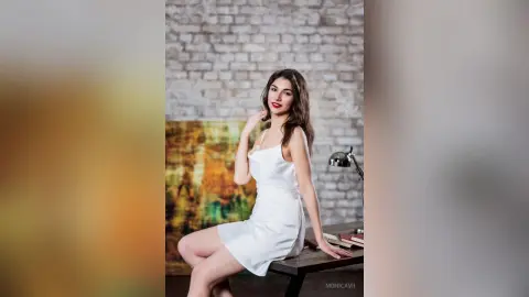 Elegant woman in a white satin slip dress sitting by a desk in a rustic brick studio setting.