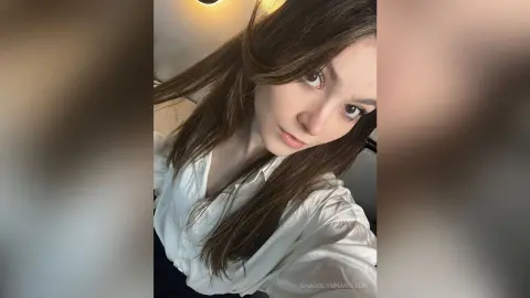Young woman with long brown hair wearing a white button-down shirt in a bright, natural indoor setting.