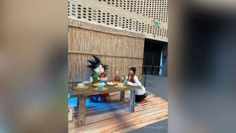 Woman in white sweater dining at a rustic wooden table next to a large Goku anime figure in a cozy cafe.