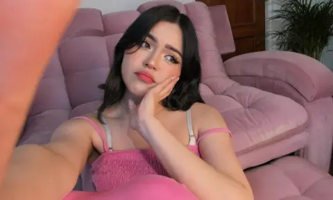 Young woman in a pink camisole relaxing on a plush pink velvet sofa with a dreamy, moody expression.
