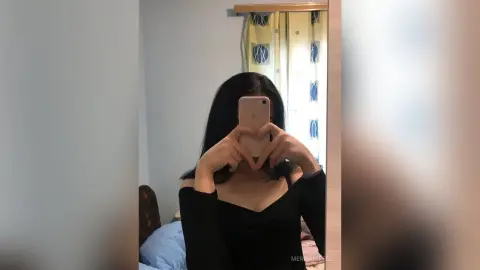 Young woman in a black long-sleeve top taking a moody mirror selfie in a bright bedroom.
