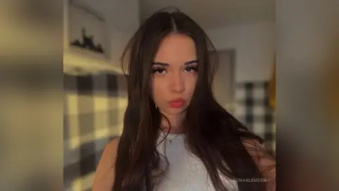 Young woman with long dark hair wearing a white tank top in a warm, sunlit indoor setting.