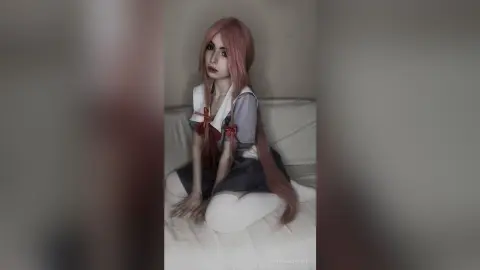 Cosplayer with long pink hair wearing a school uniform sitting on a white sofa in a soft, moody setting.