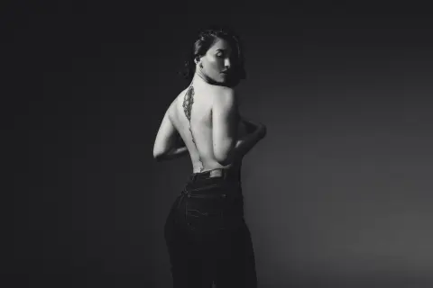 Katt_Henao poses with her back turned, showing her bare back and tattoo, wearing dark high-waisted jeans against a dark background.