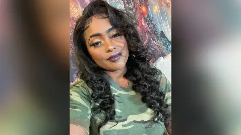 BossyBella — BossyBella poses with long, wavy black hair and dark lipstick, wearing a camouflage t-shirt against a colorful background.