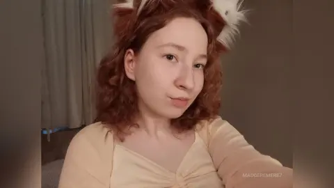 MadgeRemerez poses with her curly red hair and white headpiece, wearing a light yellow top in a room with white curtains.