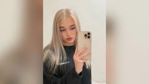 Blonde woman in a black sweatshirt taking a moody mirror selfie in a bright room.