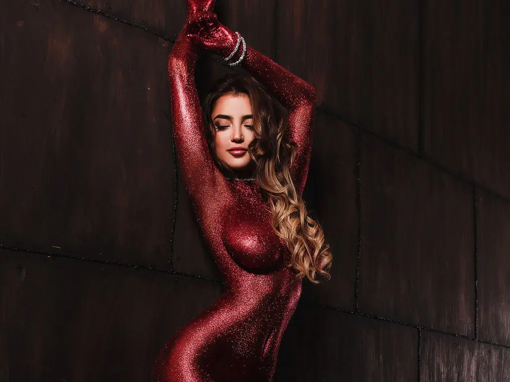 Glamorous woman in a sparkling red bodysuit posing against a dark textured wall in moody studio lighting.