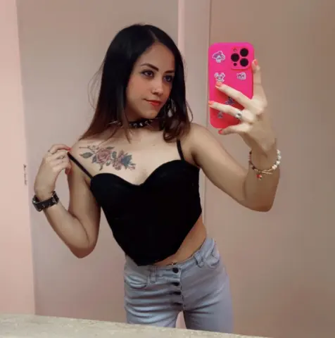 Young woman in a black corset top and light denim jeans taking a mirror selfie in a bright room.