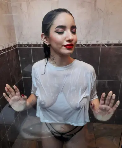 Young woman with wet hair and red lipstick wearing a white t-shirt in a tiled shower setting.