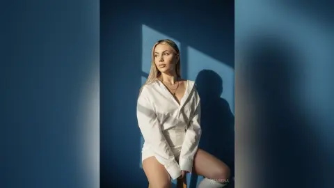 Blonde woman in a white button-down shirt posing against a blue wall in dramatic sunlight.