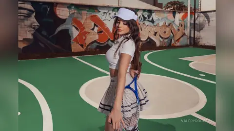 Young woman in white tennis outfit and cap posing on a green court with colorful graffiti background.