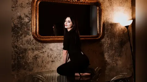 Elegant woman in a black outfit posing in front of a vintage gold mirror in a moody, rustic room.