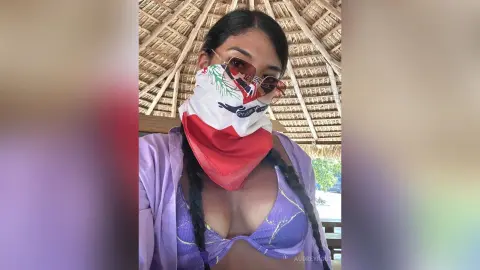 Young woman in a purple bikini and patterned bandana wearing sunglasses under a thatched roof in a sunny setting.