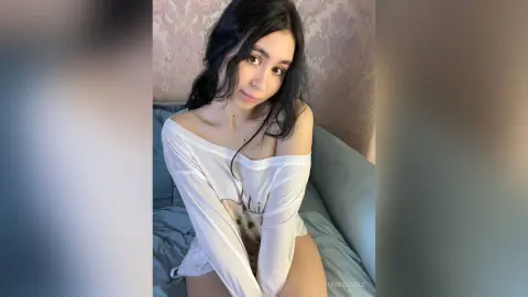 MinnieMile poses on a couch wearing a white long-sleeve shirt that hangs off her shoulders, showing her bare chest and stomach.