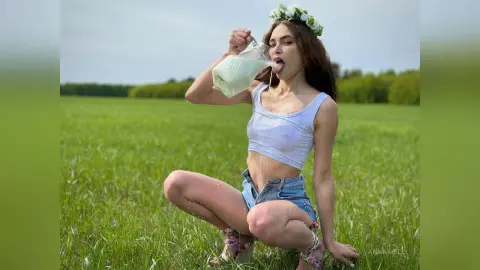 Young woman in white tank top and denim shorts drinking milk in a sunny green meadow.