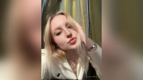Blonde woman in a white zip-up sweater posing with a soft, dreamy expression against gold curtains.