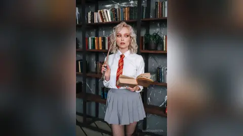 Blonde woman in white shirt and grey pleated skirt reading a book in a moody library setting.