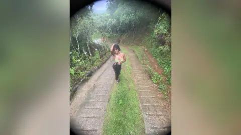 Young woman in a green bikini top and black pants walking on a forest path under a dreamy, soft light.