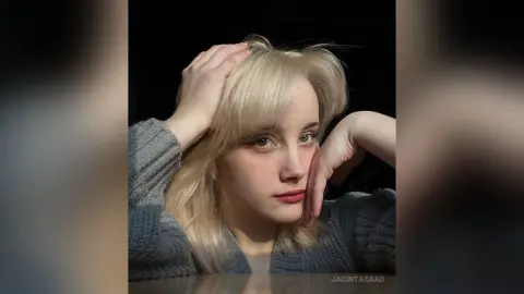 Blonde woman in a grey knit sweater posing thoughtfully against a dark background.