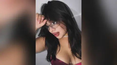 MadisonCarie with long dark hair and glasses, wearing a red top and a pearl necklace, looking down with her mouth slightly open.