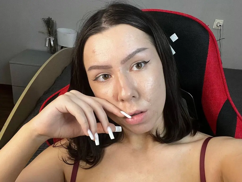 Young woman with long dark hair and winged eyeliner wearing a maroon camisole in a gaming chair setup.