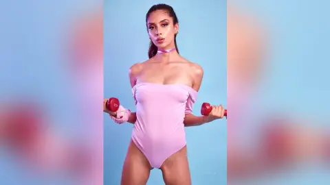 LucyMartin poses in a light pink off-the-shoulder bodysuit while holding red dumbbells against a blue background.
