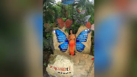 NohomiCambel stands naked in front of a large, colorful butterfly sculpture surrounded by tropical green foliage.