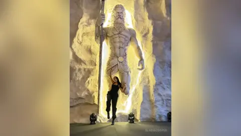 Woman in black outfit standing before a massive glowing stone statue of a bearded mythological god in a cave.
