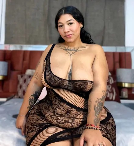 NatashaSantt — NatashaSantt wears a black lace sheer dress while sitting on a bed, showing her cleavage and tattoos.