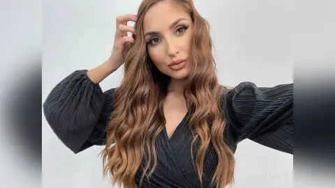 Beautiful woman with long wavy hair wearing a stylish black pleated long-sleeve dress against a white background.