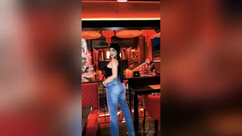 Stylish woman in black tank top and blue jeans posing in a moody, red-lit lounge with decorative lanterns.