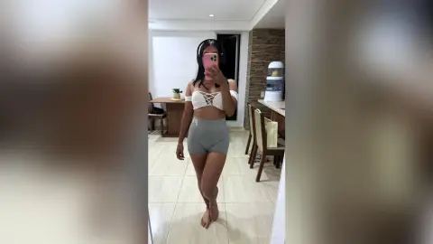Young woman in white lace crop top and grey yoga shorts taking a mirror selfie in a bright modern living room.