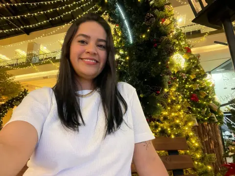 Smiling woman in a white t-shirt sitting by a glowing Christmas tree in a festive indoor setting.