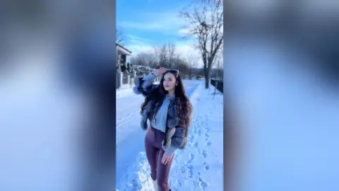 Young woman in a grey sweater and brown leather pants smiling in a snowy winter landscape under a blue sky.