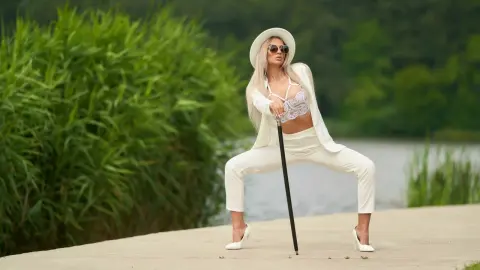 Anastasia poses outdoors in white pants and a white lace bra, wearing a white hat and sunglasses while leaning on a black cane.
