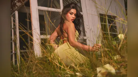 Young woman in a yellow sundress sitting in tall grass by a rustic white house during golden hour.