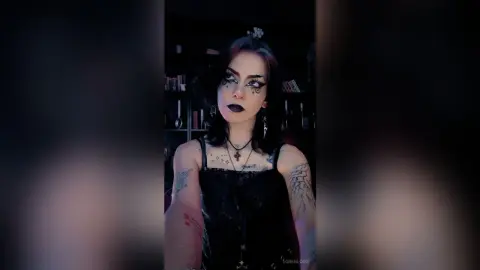 Goth woman with dark makeup and black lace outfit posing in a moody, dimly lit room.