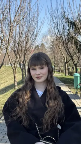 AshleyFierce poses outdoors with long wavy hair and bangs, wearing a black top and a silver chain necklace against a backdrop of bare trees.