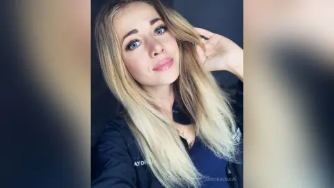 Blonde woman with blue eyes wearing a black jacket in a soft, natural light portrait.