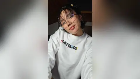 Smiling young woman with glasses wearing a white graphic sweatshirt in a cozy indoor setting.