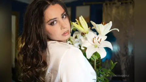 Elegant woman in a white silk blouse posing with white lilies in a moody, dimly lit indoor setting.