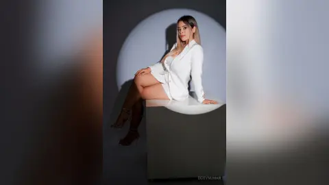 Elegant woman in a white blazer and shorts sitting on a pedestal in a minimalist studio setting.