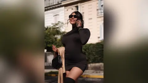 Smiling woman in a black long-sleeve mini dress and sunglasses walking outdoors on a sunny day.