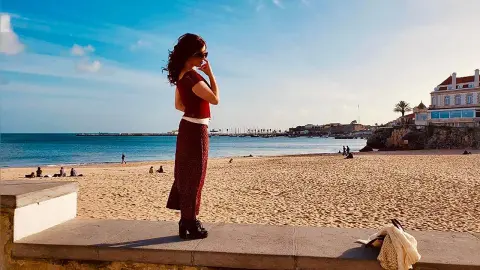 Woman in a red top and patterned skirt standing on a beach wall overlooking the ocean under a blue sky.