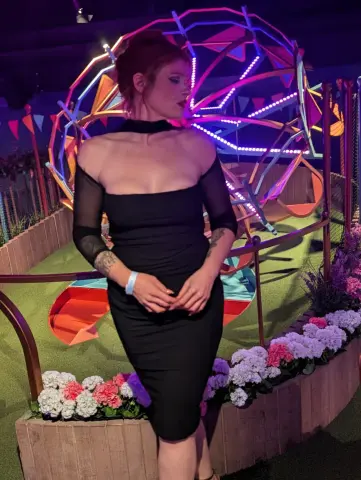 Woman in a black cocktail dress posing in front of a glowing neon geometric installation at a night event.