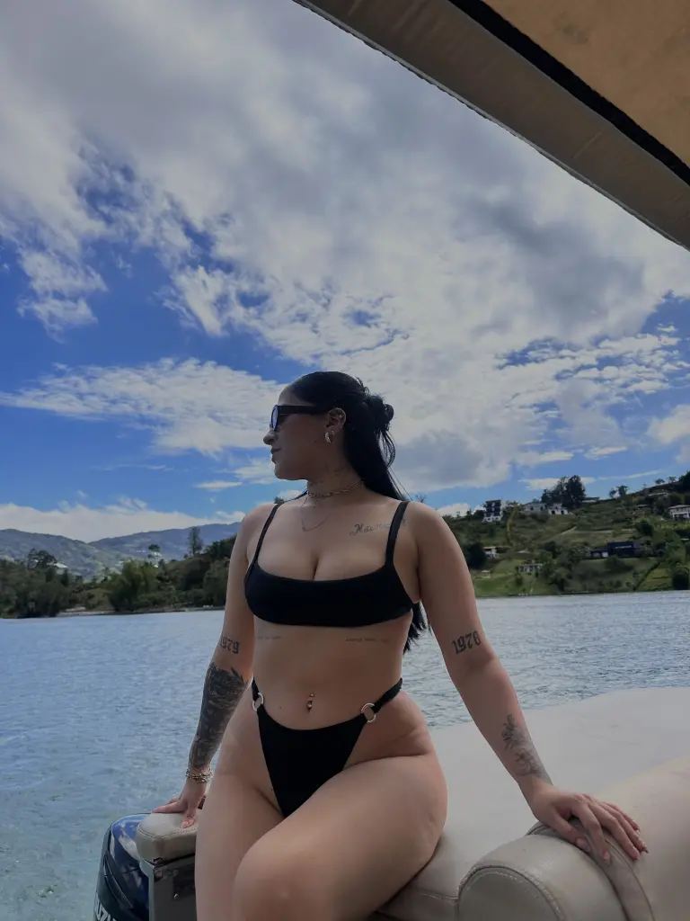 Kaiiri poses naked from the waist up, wearing a black halter bikini top and a striped beanie, looking directly at the camera in her bedroom.