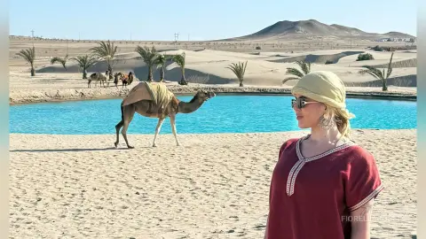 Smiling woman in a red tunic and yellow headscarf watching a camel walk near a desert oasis pool.