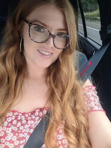 Anna_OG smiling in a car wearing glasses and a red floral top, with her long red hair falling over her shoulders.
